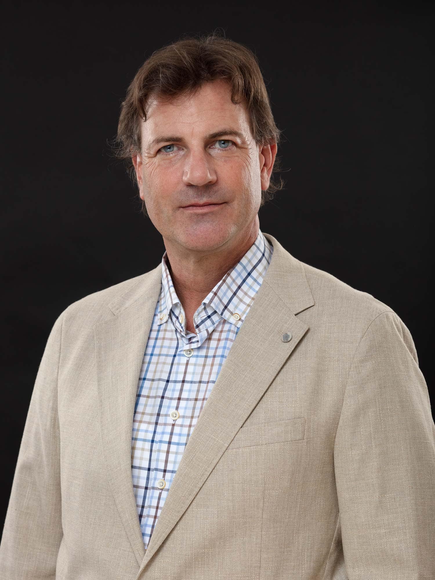 Ein Mann mit braunen Haaren, der einen hellbeigen Blazer über einem blau-weiß karierten Hemd trägt, steht vor einem schlichten dunklen Hintergrund und blickt mit neutralem Blick direkt in die Kamera - perfekt, um für die Vermietung von Lebensräumen zu werben.