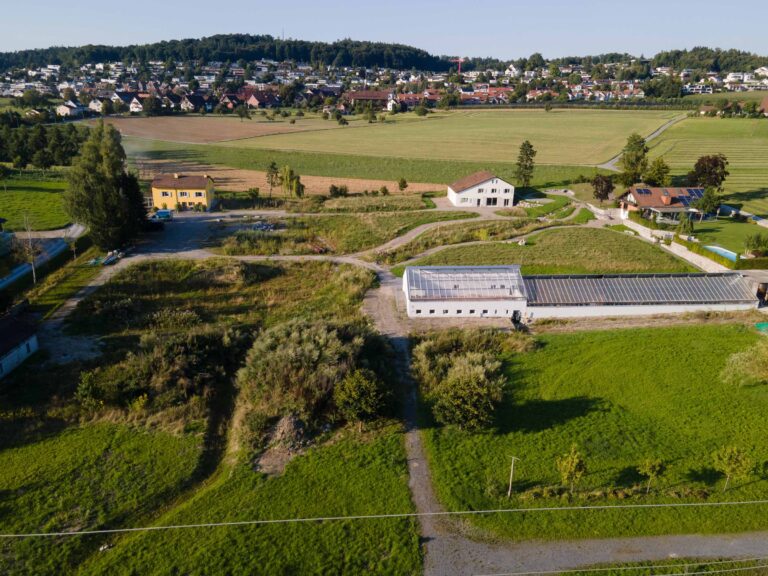 Luftaufnahme einer ländlichen Gegend mit grünen Feldern, verstreuten Häusern und einem Gewächshaus. Das kleine Dorf im Hintergrund zeigt verschiedene Lebensräume, umgeben von Bäumen und Hügeln unter einem klaren Himmel.