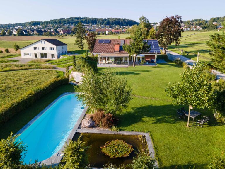 Luftaufnahme eines Hauses mit Sonnenkollektoren, großer grüner Rasenfläche, Swimmingpool, Teich und Bäumen in ländlicher Umgebung - ideal für Verwaltung oder Vermietung, mit einem weiteren Haus und Feldern im Hintergrund.