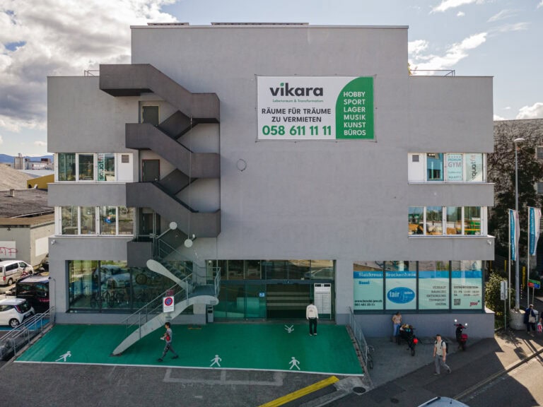 Ein modernes graues Gebäude am Leimgrubenweg 9 in Basel, mit Außentreppen und großen Fenstern. Auf der Grünfläche vor dem Gebäude parken Fahrräder und gehen Menschen spazieren. Schilder werben für die Vermietung von Hobby- und Büroräumen, auf denen eine große Kontaktnummer angegeben ist.