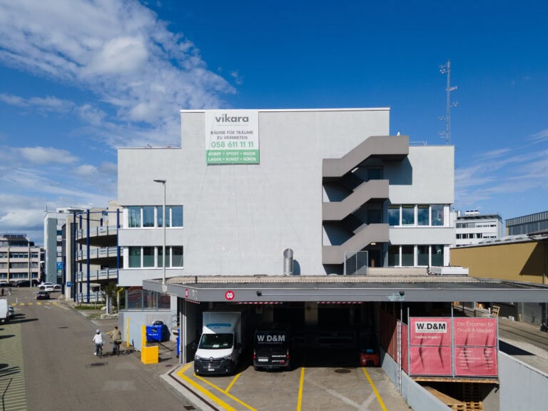 Ein modernes weißes Bürogebäude am Leimgrubenweg 9 in Basel mit grauer Außentreppe, großen Fenstern und einem grünen 