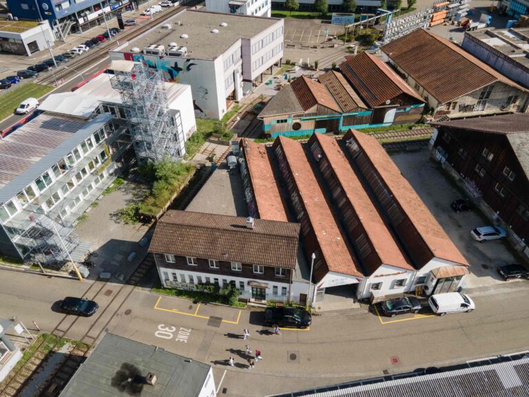 Luftaufnahme eines Industriegebiets mit langen, rot gedeckten Gebäuden, weißen Verwaltungsbüros, geparkten Autos und Menschen, die die mit einer Geschwindigkeitsbegrenzung 