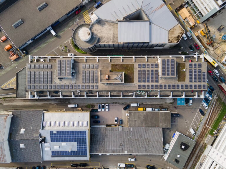 Luftaufnahme eines Industriegebäudes mit Flachdach, auf dem zahlreiche Solarzellen installiert sind, umgeben von Straßen, parkenden Autos, Verwaltungsbüros und benachbarten Gewerbebauten.