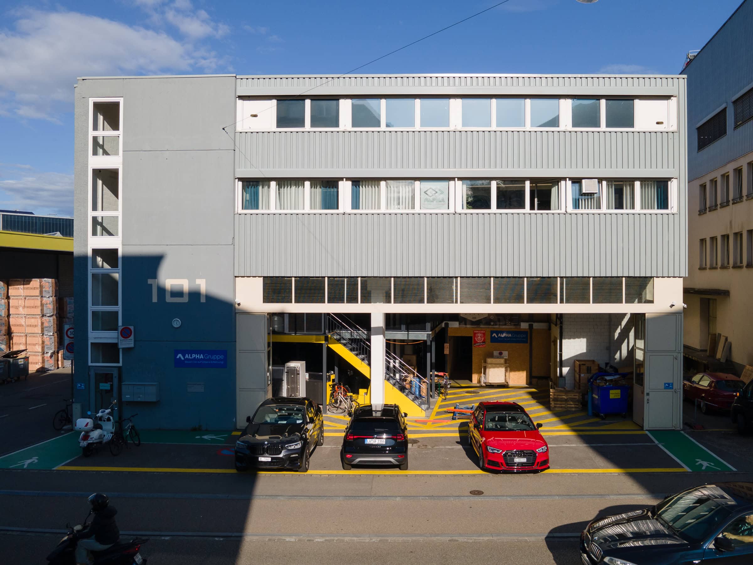 Ein modernes Industriegebäude mit großen Fenstern, beschriftet mit "T01". Davor sind drei Autos geparkt, und eine gelbe Treppe führt nach oben. Perfekt für Mietangebote und Verwaltung - der Himmel ist klar mit ein paar Wolken.
