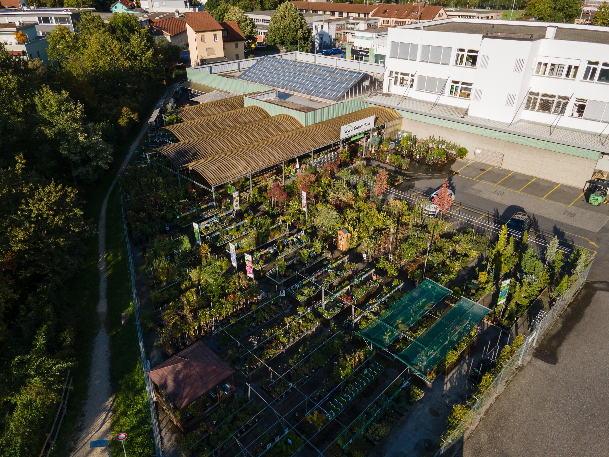 Luftaufnahme einer Gärtnerei mit Pflanzenreihen, Gewächshäusern und schattigen Bereichen; angrenzend an ein modernes weißes Verwaltungsgebäude und einen Parkplatz, mit Wohnhäusern und Bäumen im Hintergrund.