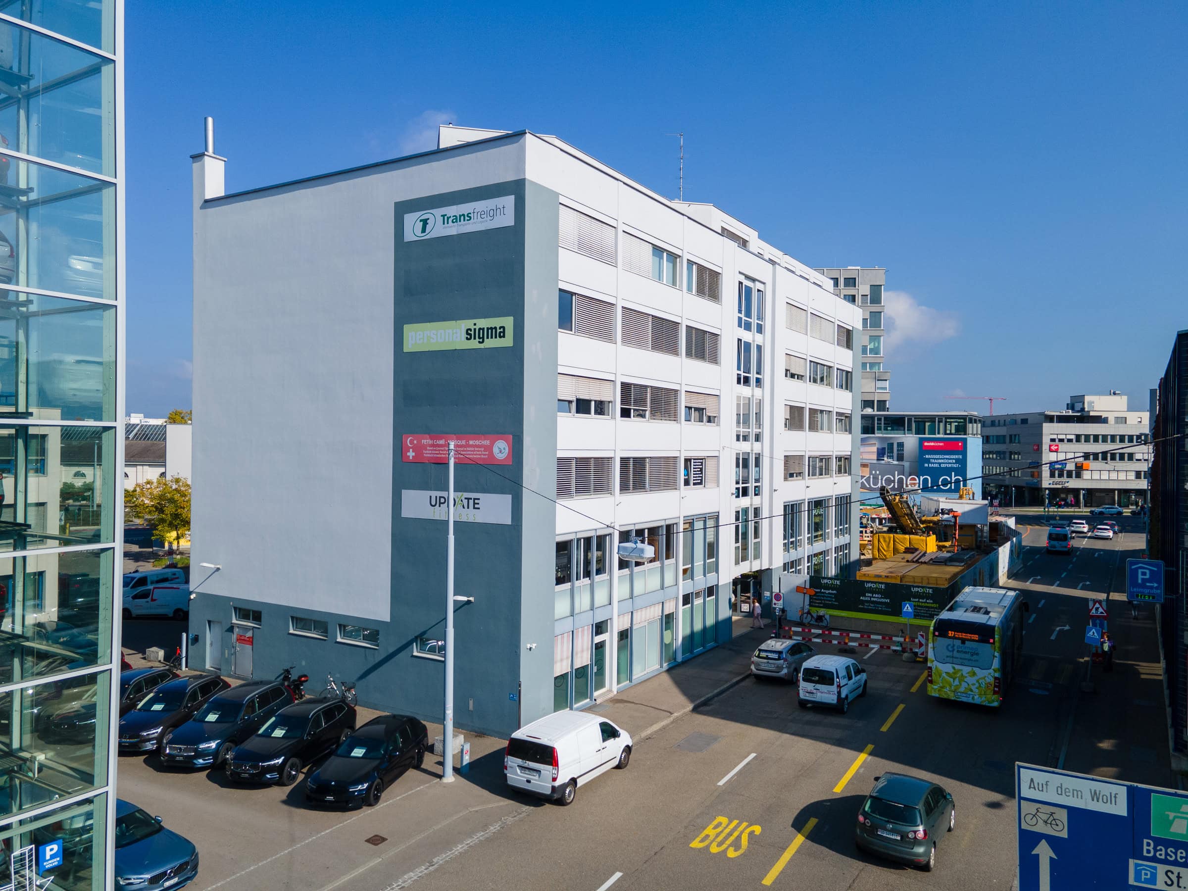 Ein weißes mehrstöckiges Bürogebäude mit Verwaltungsschildern, geparkten Autos, einem Bus an der Haltestelle und Fußgängern an einem sonnigen Tag in städtischen Lebensräumen.