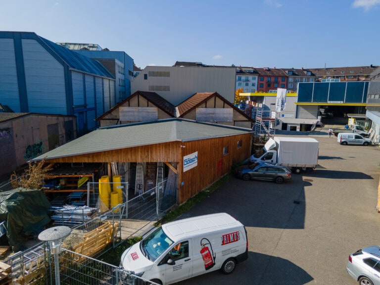 Ein hölzernes Industriegebäude mit begrüntem Dach ist umgeben von Lieferwagen und einem Lastwagen auf einem gepflasterten Parkplatz. In der Nähe gibt es Metallkonstruktionen, moderne Gebäude und Wohnungsdächer - ideal für Verwaltung oder Mietangebote von Gewerbeliegenschaften.