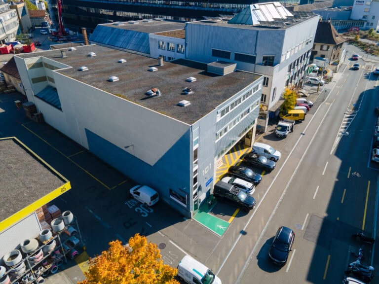 Luftaufnahme von Gewerbeliegenschaften mit einem Geschäftsgebäude mit geparkten Autos, einem gelben Kiosk und einem Fahrradweg in einem städtischen Gebiet. In der Nähe befinden sich eine Tankstelle, Herbstbäume und Flächen, die sich ideal für Verwaltung oder Vermietung eignen.