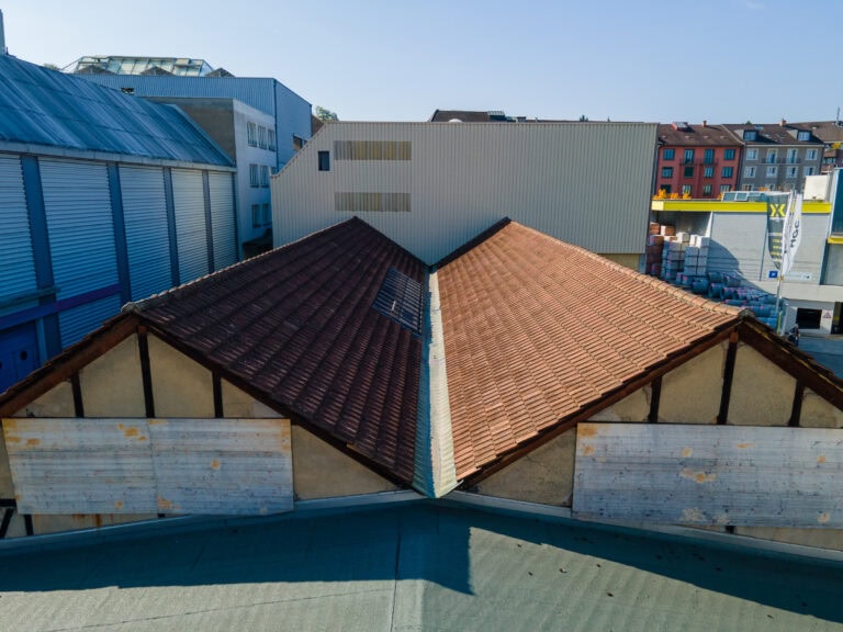 Luftaufnahme von zwei geneigten Dächern mit roten Ziegeln, die an einer zentralen Naht verbunden sind, umgeben von Verwaltungs- und Wohngebäuden, die vielfältige Lebensräume unter einem klaren blauen Himmel schaffen.