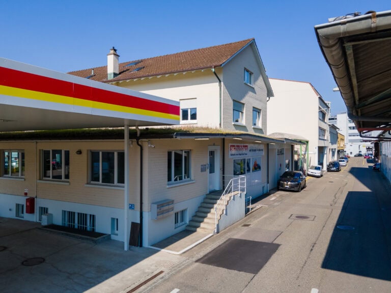 Eine Straßenansicht zeigt ein helles Gebäude mit einem Giebeldach und einer kleinen Treppe am Eingang, neben einem Tankstellenvordach - ideal für Gewerbeliegenschaften. Mehrere Autos und andere Gebäude säumen die ruhige Straße unter einem strahlend blauen Himmel.