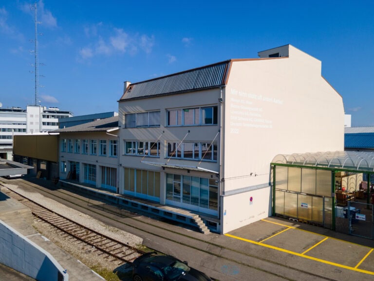 Ein modernes Industriegebäude für Gewerbeliegenschaften mit großen Fenstern und weißen Wänden, das auf einer Seite mit Text versehen ist. Davor befinden sich Eisenbahnschienen und ein Auto, über dem blauen Himmel sind Wolken zu sehen.