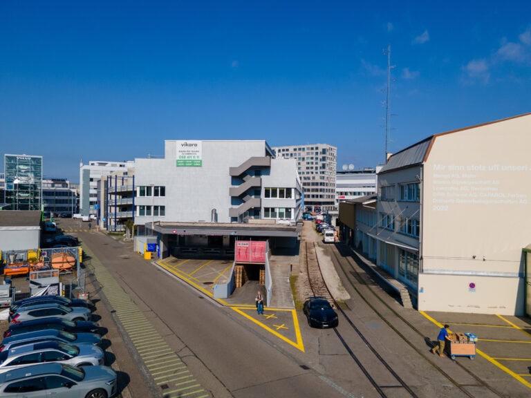 Eine Straßenszene in einem städtischen Industriegebiet mit modernen Gebäuden, geparkten Autos und einer Person, die unter einem klaren blauen Himmel auf dem Bürgersteig geht. Eine Wandmalerei mit deutschem Text hebt Gewerbeliegenschaften und Vermietung an einer Gebäudeseite hervor.