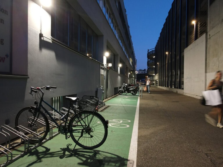 Ein schwach beleuchteter städtischer Radweg in der Abenddämmerung mit geparkten Fahrrädern auf der linken Seite, sichtbaren Mietangebotsschildern, Motorrädern in der Ferne und zwei verschwommenen Fußgängern, die den Weg zwischen hohen Gebäuden entlanggehen.