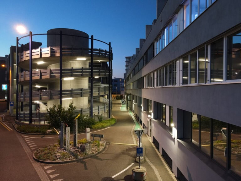 Eine spiralförmige Parkhausrampe wird in der Abenddämmerung neben einem modernen Gebäude mit großen Fenstern beleuchtet, die durchdacht gestaltete Lebensräume hervorheben, während Straßenlaternen und ein paar Schilder die ruhige, leere Straße säumen.