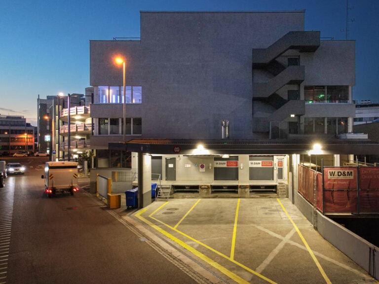 Eine Laderampe am Leimgrubenweg 9 in Basel in der Abenddämmerung, mit leeren Parkbuchten, gelben Linien auf dem Boden und einem wegfahrenden Lastwagen. Das Gebäude dahinter hat eine Außentreppe und beleuchtete Fenster, während die Straßenlaternen ein warmes Licht werfen.