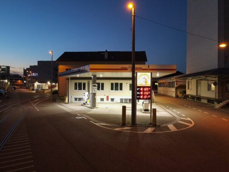 Eine kleine Tankstelle, die von einer professionellen Verwaltung geführt wird, ist in der Abenddämmerung in einer ansonsten leeren Straße hell erleuchtet. Das Preisschild leuchtet bei Einbruch der Dunkelheit unter einer Straßenlaterne, und die umliegenden Lebensräume sind schwach gegen den tiefblauen Himmel beleuchtet.