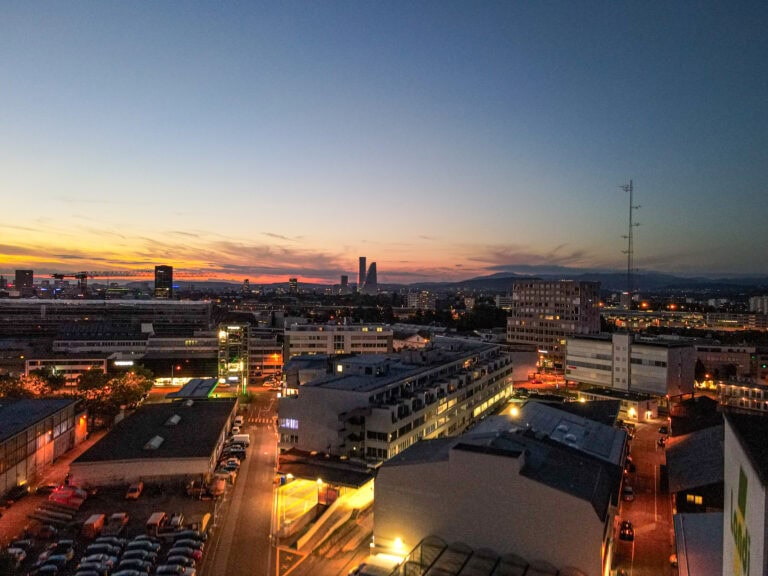 Ein Stadtbild in der Abenddämmerung in Basel, mit beleuchteten Gebäuden und Straßen am Leimgrubenweg. Der Himmel leuchtet orange und blau, während die Sonne hinter fernen Bergen und hohen Gebäuden am Horizont untergeht.
