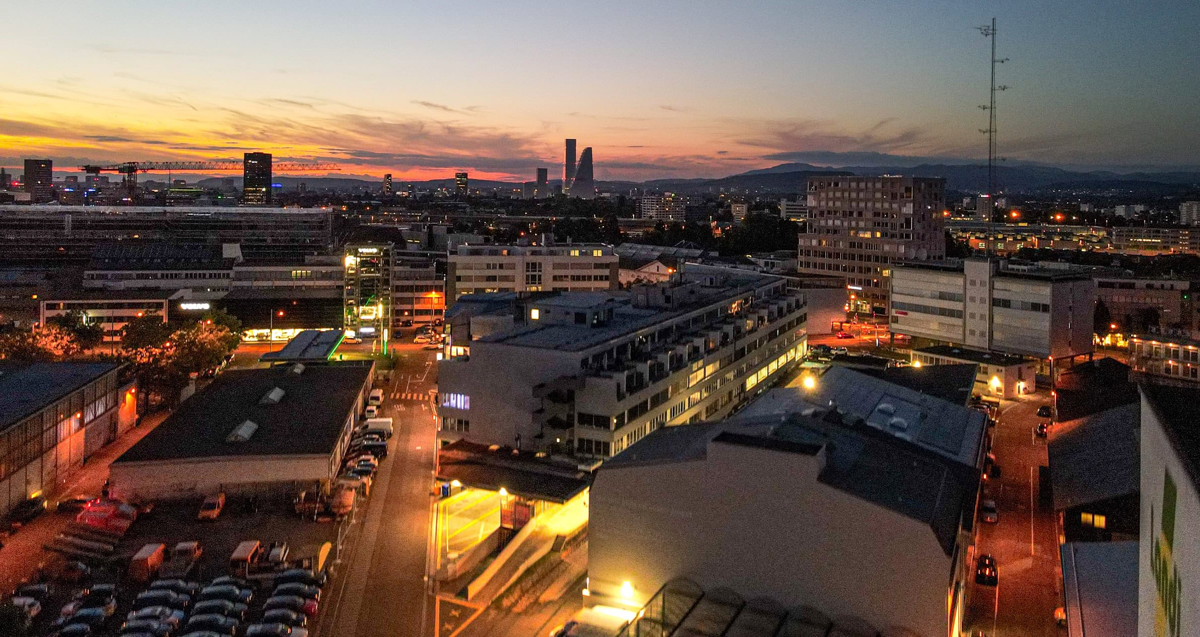 Ein Stadtbild in der Abenddämmerung in Basel mit beleuchteten Straßen, modernen Gebäuden und zwei hohen Wolkenkratzern in der Nähe des Leimgrubenwegs 9 unter einem farbenfrohen Sonnenuntergangshimmel. Am Horizont sind Berge zu sehen.