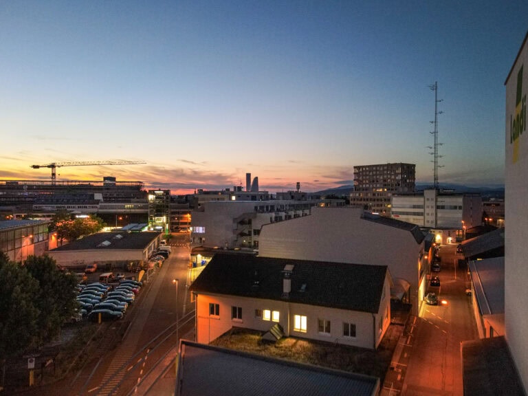Ein Stadtbild in der Abenddämmerung in Basel mit beleuchteten Gebäuden, parkenden Autos und Straßenlaternen. Der Himmel zeigt einen Farbverlauf von Orange zu Tiefblau, und in der Nähe des Leimgrubenwegs 9 erheben sich in der Ferne zwei hohe Wolkenkratzer.