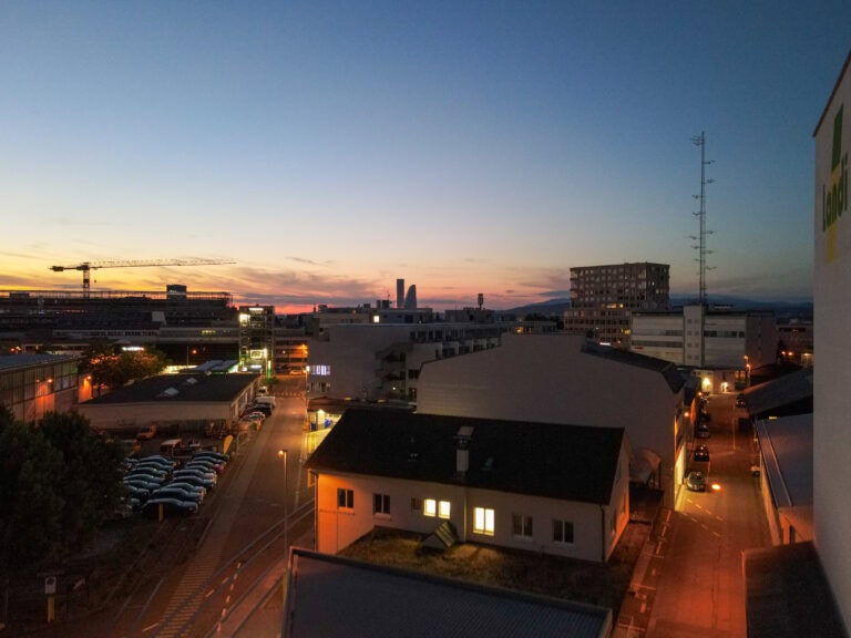 Eine Stadtlandschaft in der Abenddämmerung mit von Straßenlaternen beleuchteten Gebäuden und Straßen. Der Himmel wechselt bei Sonnenuntergang von Blau zu Orange und hebt verschiedene Lebensräume inmitten von entfernten Wolkenkratzern und Bergen am Horizont hervor.