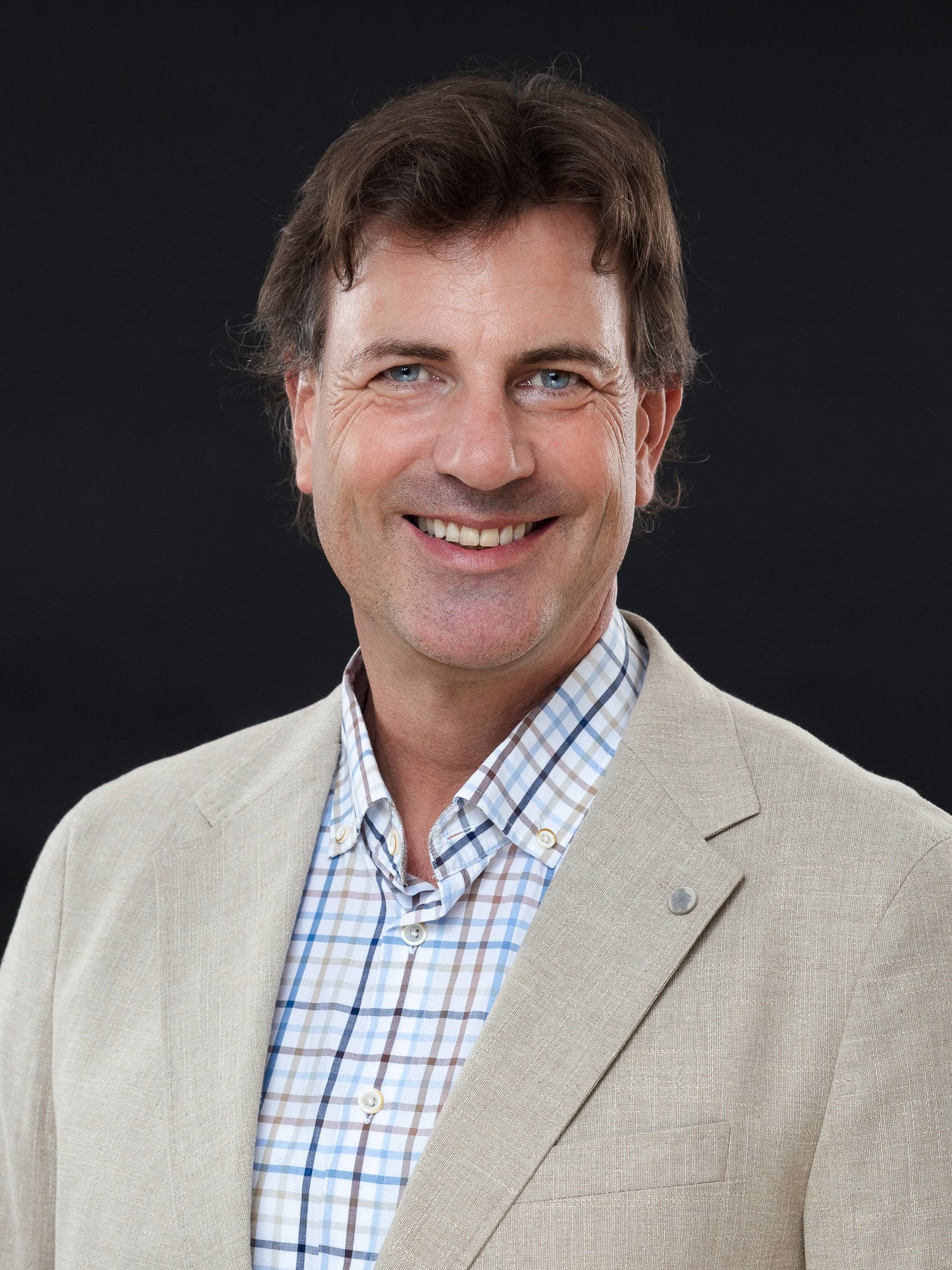 Ein Mann mit braunen Haaren, der einen beigen Blazer und ein blau-weiß kariertes Hemd trägt, lächelt vor einem schlichten schwarzen Hintergrund und ist bereit, über Mietangebote zu diskutieren und Einblicke in Lebensräume zu geben.
