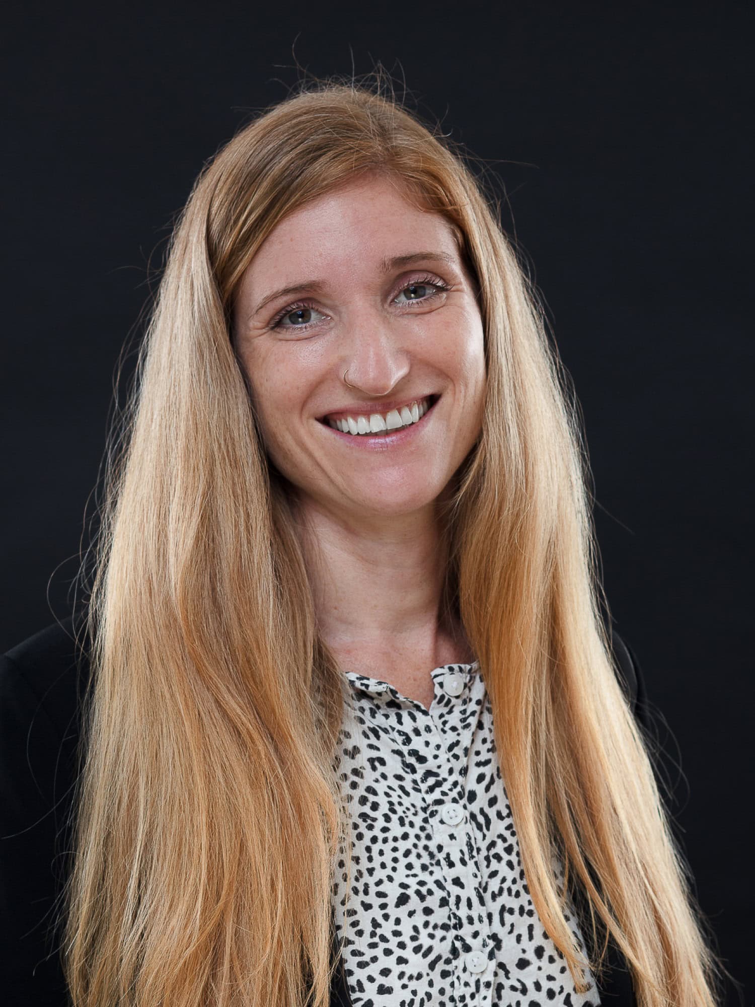 Eine Frau mit langen blonden Haaren lächelt in die Kamera. Sie trägt einen schwarzen Blazer über einer weißen Bluse mit schwarzem Tupfenmuster vor einem einfarbigen schwarzen Hintergrund - perfekt für die Präsentation von Vermietungs- oder Mietangeboten.