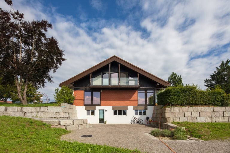 Ein modernes Haus mit schrägem Dach, großen Fenstern, roten Ziegeln und weißen Wänden. Es eignet sich perfekt für die Verwaltung oder Vermietung und ist mit Steinmauern, Grünflächen, einem teilweise bewölkten Himmel und einem neben der Tür geparkten Fahrrad ausgestattet.