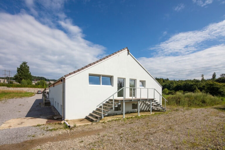 Ein weißes, einstöckiges Verwaltungsgebäude mit schrägem Dach und zwei Außentreppen, die zu separaten Eingängen führen, umgeben von Kies und Grünflächen unter einem teilweise bewölkten blauen Himmel.