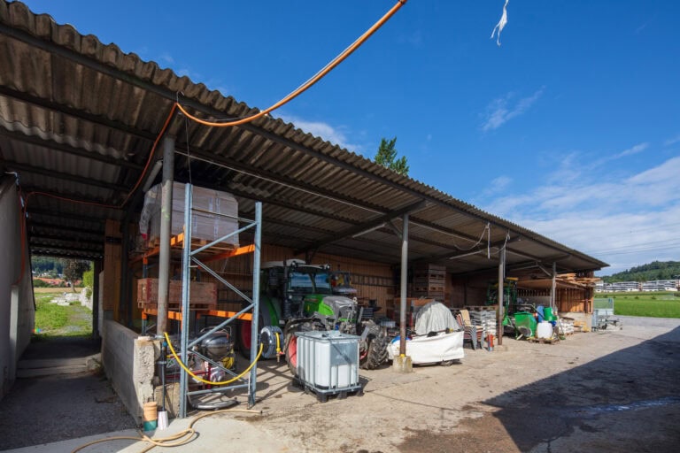 Ein landwirtschaftlicher Lagerschuppen mit einem Wellblechdach beherbergt verschiedene Geräte, Werkzeuge und Vorräte. Ideal für Gewerbeliegenschaften oder Mietangebote. Ein grüner Traktor und große Tanks sind zu sehen, mit Feldern und blauem Himmel im Hintergrund.