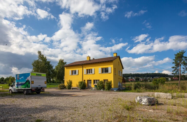 Ein leuchtend gelbes zweistöckiges Haus steht in einer ländlichen Gegend unter einem teilweise bewölkten Himmel, umgeben von offenen Feldern. In der Nähe parkt ein weiß-grüner Lieferwagen auf dem Schotterplatz - eine einladende Szene für Mietangebote und neue Lebensräume.
