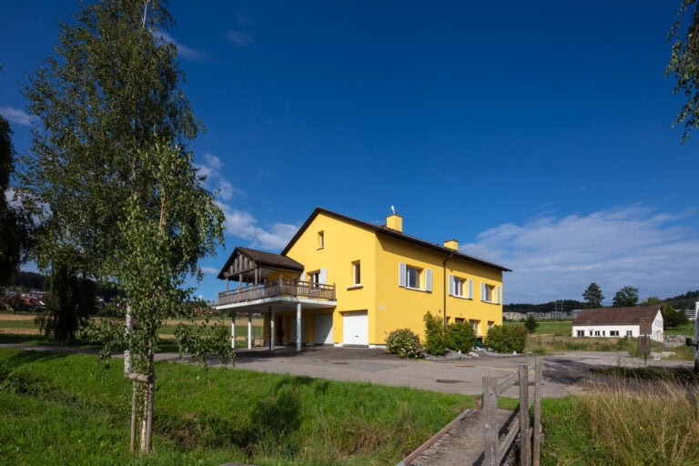 Ein leuchtend gelbes, zweistöckiges Haus mit weißen Verkleidungen steht in einer ländlichen Landschaft, umgeben von grünem Gras und Bäumen unter einem klaren blauen Himmel, der auf Verwaltung oder mögliche Mietangebote hindeutet, wobei im Hintergrund ein weiteres kleines Gebäude zu sehen ist.