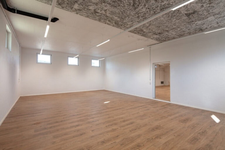 Ein geräumiger, leerer Raum mit weißen Wänden, einem holzgemusterten Fußboden, vier hohen quadratischen Fenstern - ideale Lebensräume für die Vermietung von Gewerbeliegenschaften - mit Leuchtstoffröhren an der Decke und einer offenen Tür, die in einen anderen Raum führt.