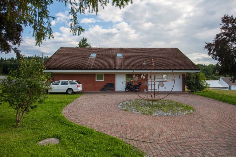 Ein Haus mit braunem Schrägdach, weißen Ziegelwänden und einer kreisförmigen Einfahrt mit einer Metallskulptur. Perfekt für Verwaltung oder Vermietung, mit Mietangeboten verfügbar. Ein weißes Auto und ein Motorrad stehen in der Nähe der Garage; Bäume und Gras umgeben das Haus.