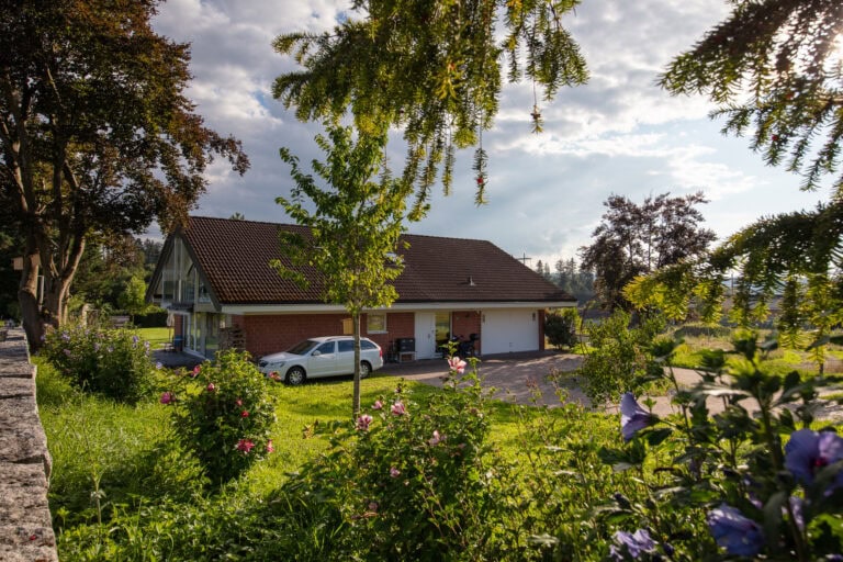 Ein Haus mit rotem Schrägdach und Garage steht inmitten von grünem Gras, blühenden Sträuchern und Bäumen unter einem teilweise bewölkten Himmel. In der Einfahrt parkt ein weißes Auto - ideal für Vermieter oder diejenigen, die Mietangebote für neue Lebensräume suchen.