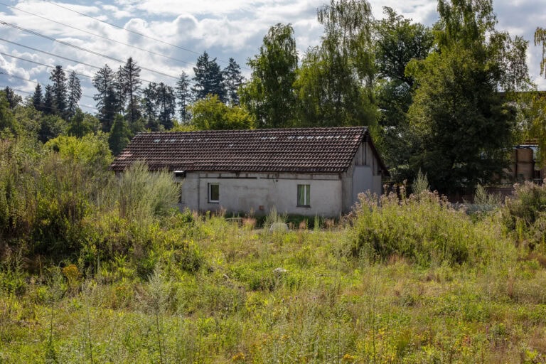 Ein kleines, weißes, einstöckiges Haus mit rotem Ziegeldach steht auf einer bewachsenen Wiese, umgeben von grünen Bäumen - ein einladender Ort inmitten der Natur, ideal für Verwaltung oder einzigartige Mietangebote unter einem teilweise bewölkten Himmel.