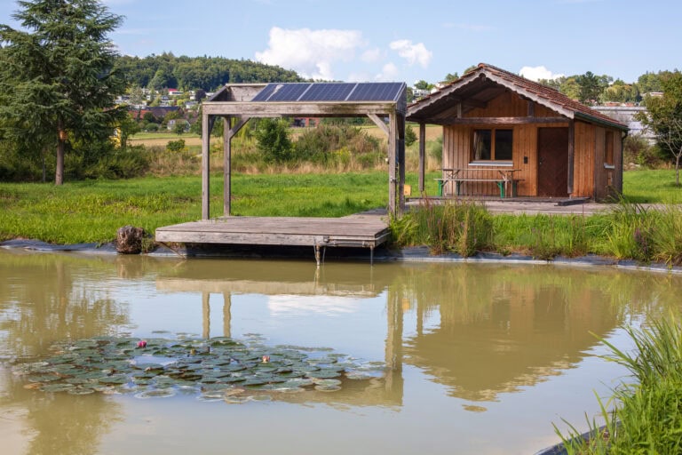 Eine kleine Holzhütte mit einer überdachten Veranda steht neben einem Teich mit Seerosen, eingebettet in grasbewachsene Lebensräume und umgeben von Bäumen, mit ein paar Häusern im fernen Hintergrund unter blauem Himmel.