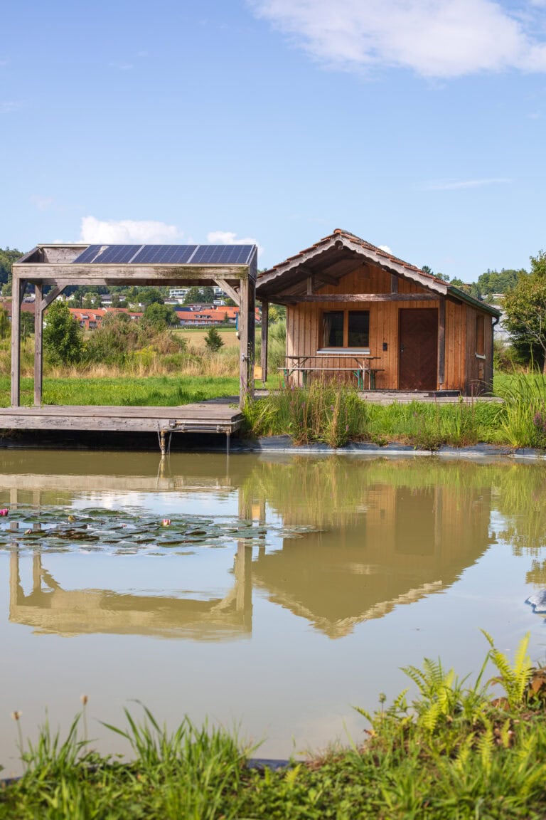 Eine kleine Holzhütte und ein überdachter Steg liegen neben einem Teich mit Seerosen, die sich auf dem ruhigen Wasser spiegeln. Diese heitere Szene ist von üppigem Grün umgeben und bietet inspirierende Lebensräume unter einem blauen Himmel mit einigen Wolken.