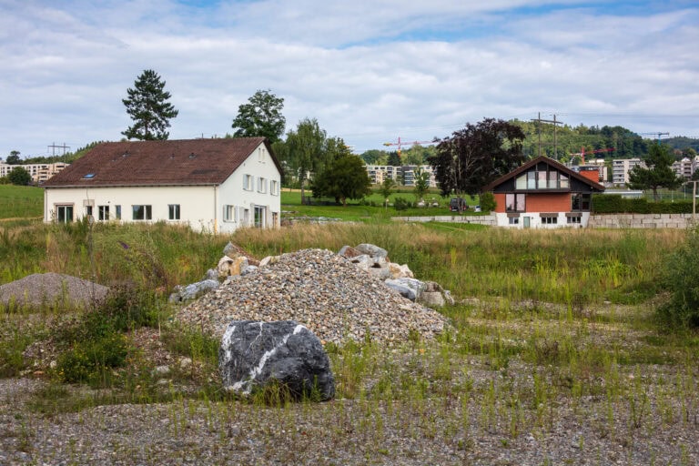 Ein grasbewachsenes Feld mit Schotter- und Steinhaufen im Vordergrund, zwei Häuser im Mittelgrund und Wohnhäuser, Bäume und ein blauer Himmel mit Wolken im Hintergrund - eine sich entwickelnde Szene zwischen Lebensräumen und Gewerbeliegenschaften.
