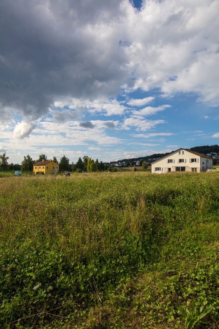 Ein weites, grasbewachsenes Feld erstreckt sich in Richtung von zwei Häusern, einem gelben und einem weißen - möglicherweise für Verwaltung oder Vermietung - unter einem teilweise bewölkten blauen Himmel. Im Hintergrund sind Bäume und entfernte Hügel zu sehen.