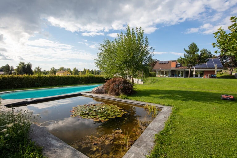 Ein Hinterhof-Lebensraum mit einem kleinen Teich mit Seerosen, einem rechteckigen Swimmingpool, grünem Rasen und einem Haus mit Sonnenkollektoren unter einem teilweise bewölkten Himmel. Auf dem Rasen ist ein Rasenmähroboter zu sehen.