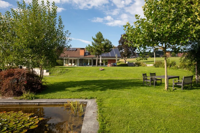 Ein grasbewachsener Hinterhof mit Bäumen, einem Teich im Vordergrund und einem Tisch mit Stühlen. Im Hintergrund ist ein Haus mit Sonnenkollektoren auf dem Dach zu sehen - perfekt für Vermietung oder Mietangebote -, umgeben von Grün unter einem teilweise bewölkten Himmel.