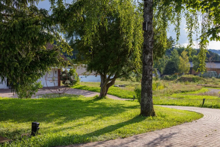 Ein sonnenbeschienener Garten mit grünem Gras, hohen Bäumen und einem gewundenen Ziegelweg schafft harmonische Lebensräume. Im Hintergrund erscheinen ein weißes Gebäude und ein dekorativer Metallwagen zwischen üppigem Laub und entfernten Häusern.