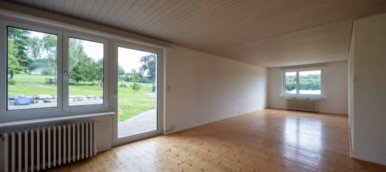 Heller, leerer Raum mit hellem Holzboden und weißen Wänden, ideal für vielfältige Lebensräume. Große Fenster und eine Glastür bieten Blick ins Grüne; perfekt für private Nutzung oder Vermietung durch professionelle Verwaltung.