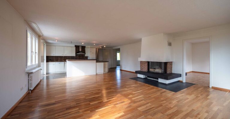 Ein geräumiges, leeres Wohnzimmer mit Holzböden, eine moderne weiße Küche im Hintergrund, große Fenster und ein zentraler Kamin mit einem gemauerten Sockel - perfekte Lebensräume, die durch unsere Mietangebote angeboten werden.