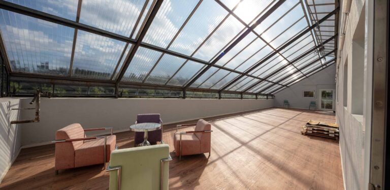 Ein geräumiger Raum mit einem schrägen Glasdach, perfekt für die Vermietung oder Verwaltung von Gewerbeliegenschaften. Das Sonnenlicht strömt herein, drei Sessel und ein kleiner runder Tisch stehen auf dem Holzboden, in der Ecke stapeln sich Paletten, und draußen sind grüne Hügel zu sehen.