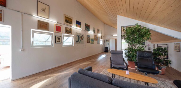 Ein modernes Loft mit Holzböden und einer schrägen Holzdecke, ideal für die Vermietung, mit einer Sitzecke mit grauen und schwarzen Stühlen, Pflanzen und Wänden mit gerahmten Kunstwerken und Fenstern, die natürliches Licht hereinlassen.