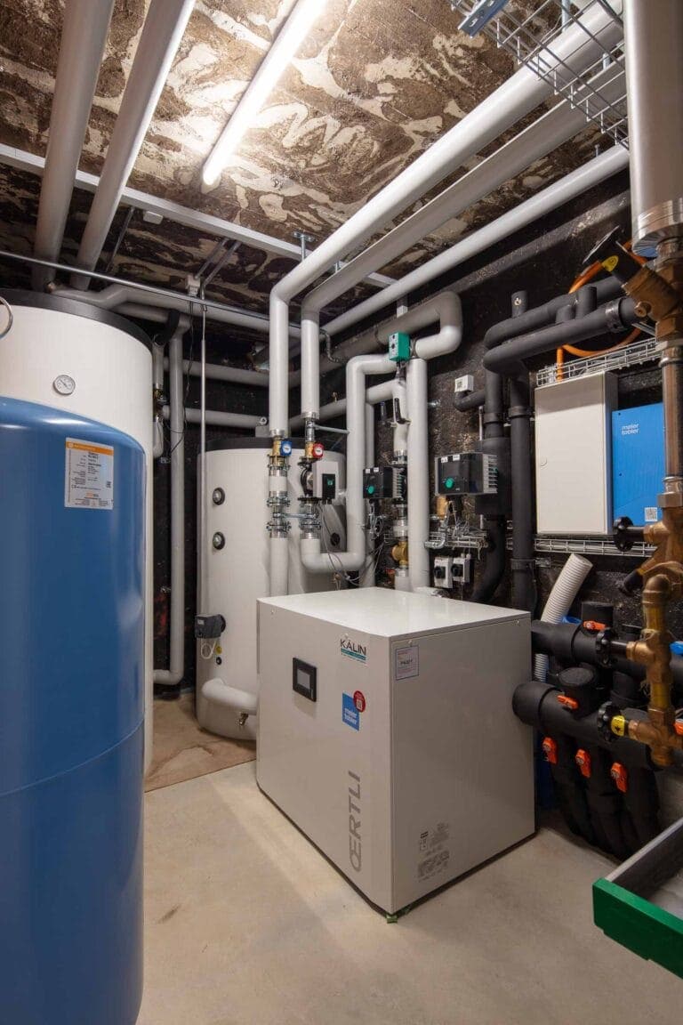 Ein sauberer, organisierter Hauswirtschaftsraum in einer Gewerbeliegenschaft mit einer weißen Wärmepumpe, einem blauen Speichertank, Rohren, Messgeräten, Ventilen und Schalttafeln, die an Betonwänden und -decken montiert sind. Eine helle Deckenbeleuchtung erhellt den Raum.