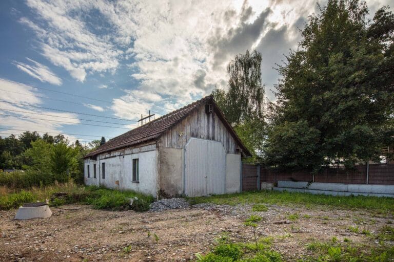 Ein alter, verwitterter weißer Schuppen mit Schrägdach steht auf einem lückenhaften, überwucherten Hof unter einem teilweise bewölkten Himmel, umgeben von Bäumen und einem Holzzaun - ein Echo vergessener Lebensräume inmitten der stillen Verwaltung der Natur.