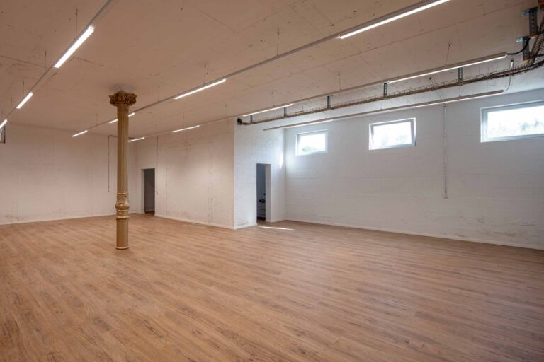 Ein geräumiger, leerer Raum mit hellem Holzfußboden, weißen Wänden, drei hohen rechteckigen Fenstern, fluoreszierenden Deckenleuchten, einer einzelnen gelben Säule und mehreren offenen Türen - ideal für die Verwaltung oder flexible Mietangebote.