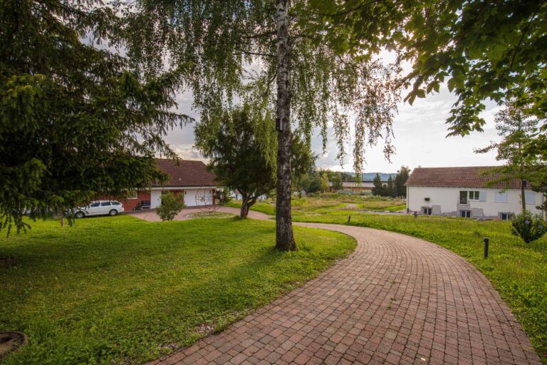 Ein geschwungener Ziegelsteinweg schlängelt sich durch grasbewachsene, von Bäumen gesäumte Lebensräume und führt zu Häusern mit schrägen Dächern unter einem teilweise bewölkten Himmel. Ein weißes Auto ist neben einem der Häuser geparkt.